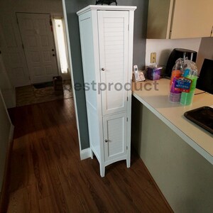 May include: A tall, white, freestanding storage cabinet featuring two doors with horizontal louvered slats and small, round, silver-toned metal knobs. The cabinet has a rectangular top surface and stands on four short, straight legs.