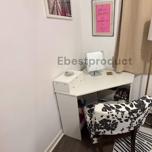 May include: A white corner vanity desk with a triangular top, featuring a white jewelry box with two drawers and a square vanity mirror on top. A chair with a black and white cow print pattern is positioned in front of the desk.