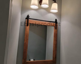 May include: A rectangular wall mirror with a medium-brown wood frame, featuring a black metal sliding barn door hardware rail at the top. Above the mirror is a three-light vanity fixture with white glass shades and a dark metal finish.