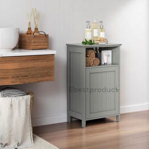 May include: A gray freestanding bathroom cabinet with a single door and an open shelf. The cabinet has a wood grain texture and is sitting on a wood floor. The cabinet is filled with toiletries and towels.