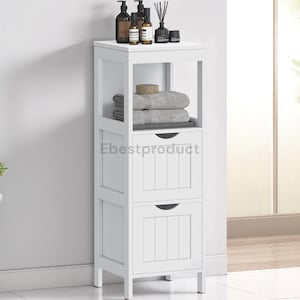 May include: A white wooden bathroom storage cabinet with two drawers and an open shelf. The top shelf holds bottles, a diffuser, and a small black container. The open shelf holds folded gray towels and a bar of soap. The cabinet is against a white wall.