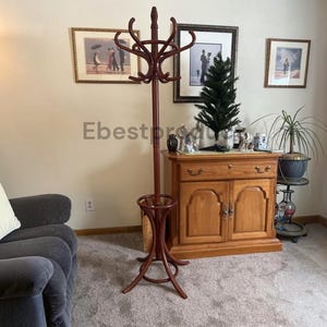 May include: A tall, dark brown wooden coat rack featuring a central pole with multiple curved hooks at the top for hanging garments and a circular base with a built-in umbrella stand at the bottom, standing on a carpeted floor.
