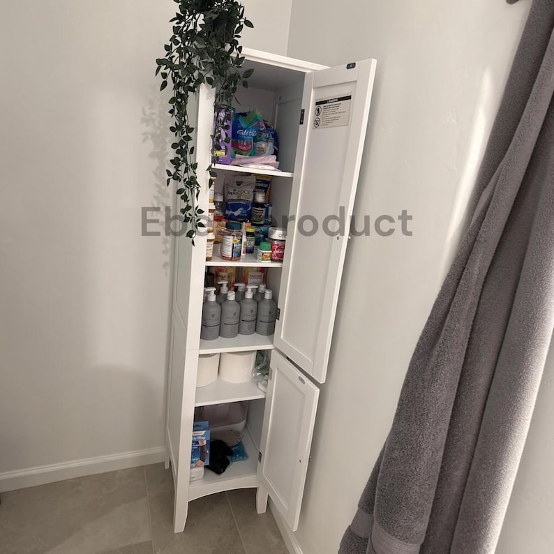 May include: A tall, white, narrow bathroom storage cabinet with two hinged doors, featuring multiple interior shelves filled with various bottles, containers, and toiletries. A green trailing plant sits on top of the unit.