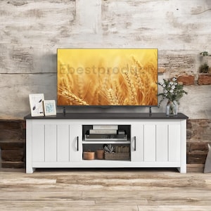 May include: White TV stand with two doors and a shelf. The stand has black hardware and a black top. The stand is in front of a brick wall with a TV on top.