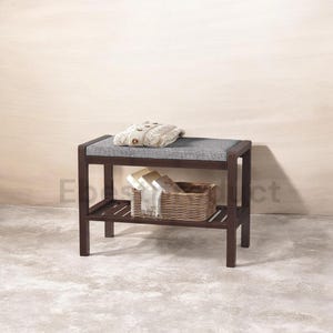 May include: A dark brown wooden bench with a gray upholstered seat. A woven basket with folded towels and a sweater are on the bench. The bench has a lower shelf for storage.