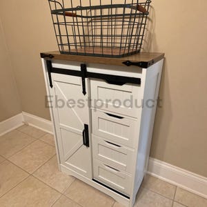 May include: A white storage cabinet with a sliding barn door, four drawers, and a brown wooden top. A black metal wire basket with wooden handles sits on the top surface. The cabinet features black metal hardware and a sliding rail mechanism.