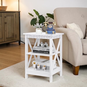May include: A white, three-tiered side table with an X-frame design. The table holds books, decorative perfume bottles, and astronaut figurines. A vintage-style telephone and a small vase with flowers sit on top. The text "Ebestproduct" is visible on the top shelf.