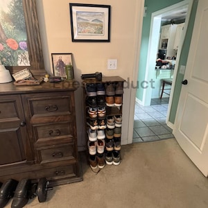 May include: A tall, narrow, dark brown wooden shoe rack with multiple shelves holding various pairs of sneakers and casual shoes. The rack is positioned against a wall next to a large wooden dresser with drawers and a black handheld massage gun on top.