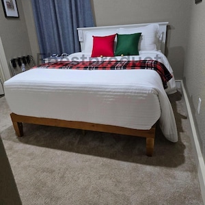 May include: A wooden bed frame with a white headboard, topped with a white quilted comforter, a red and black plaid runner, and two square throw pillows in solid red and dark green, positioned against a wall with blue curtains.
