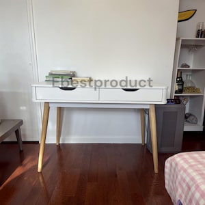 May include: A white desk with two drawers featuring black recessed handles and four light-toned wooden legs. A stack of books sits on the left side of the desk surface. The text Ebestproduct is overlaid across the center of the desk.