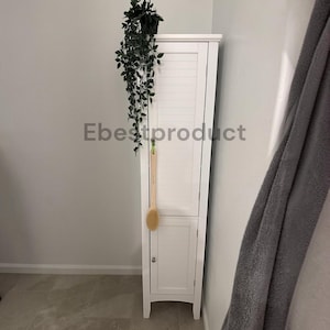 May include: A tall, white, narrow storage cabinet with two louvered doors and a small silver knob. A long-handled wooden bath brush hangs from the upper door, and a trailing green artificial plant sits on the top surface of the cabinet.
