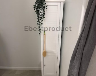 May include: A tall, white, narrow storage cabinet with two louvered doors and a small silver knob. A long-handled wooden bath brush hangs from the upper door, and a trailing green artificial plant sits on the top surface of the cabinet.