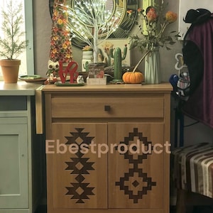 May include: A light brown wooden cabinet with one drawer and two doors, each featuring a dark brown geometric pattern. The cabinet top is decorated with a light-up tree, a red "LOVE" sign, a small cactus, a pumpkin, and a vase of flowers.