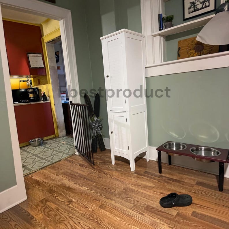 May include: A tall white storage cabinet with two louvered doors stands on a wooden floor. Next to it is a raised pet feeding station with two stainless steel bowls. A black clog shoe sits on the floor nearby, and a black metal gate is visible.
