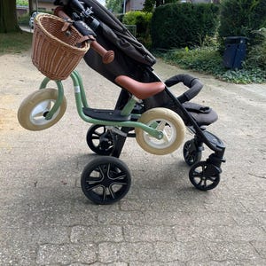 Könnte beinhalten: Ein grünes und schwarzes Kinderdreirad mit einem braunen Weidenkorb am Lenker. Das Dreirad steht neben einem schwarzen Kinderwagen mit einem braunen Ledersitz.