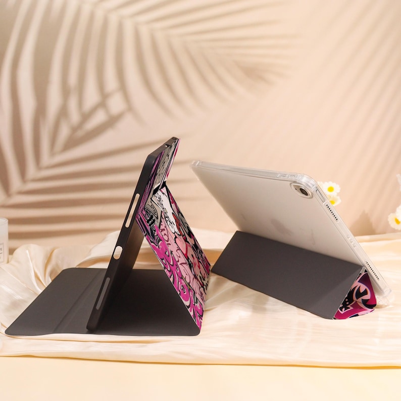 May include: Two tablet cases are shown, one open and the other closed. The open case has a bright pink and white graphic design. The closed case is black. The tablets are propped up on a stand.