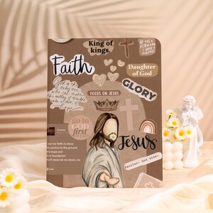 Peut inclure: Un journal marron orné d'autocollants sur le thème religieux. Les autocollants comportent les mots "Foi", "Jésus", "Gloire" et "Fille de Dieu". Un dessin de Jésus est également présent. Le journal est décoré de cœurs, de croix et de couronnes.