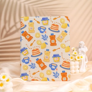May include: A beige notebook cover with a repeating pattern of honey bears, honey jars, lemons, teacups, and honey dippers. The words "Honey" and "Lemon" are also printed on the cover. The notebook is rectangular.