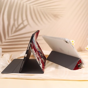 May include: Two tablet cases, one black and one clear, are displayed. The black case features a comic book design with red, white, and blue colors. The clear case is propped up with a gray stand. The background is a soft beige.
