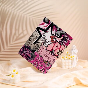 May include: A rectangular case with a graffiti design in pink, black, and white. The design incorporates a star, the Hello Kitty character, and graffiti lettering. The case is angled, displaying its colourful artwork.
