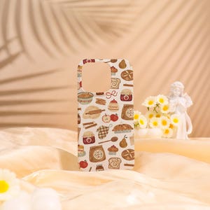 May include: A white phone case with a repeating pattern of baking-related illustrations, including apples, jars, and baked goods. The case has a cutout for the camera and buttons. The overall theme is a cozy, kitchen-inspired design.