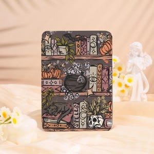May include: A decorative phone case featuring a whimsical bookshelf design. The case showcases a dark gray background with illustrations of books, pumpkins, a cauldron, and a sleeping cat. The design includes accents of pink, green, and white, creating a magical, fantasy theme.