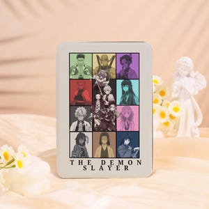 May include: A white rectangular product with a grid of colourful anime character portraits. The text "THE DEMON SLAYER" is printed in black at the bottom. The portraits are set against a pastel background.