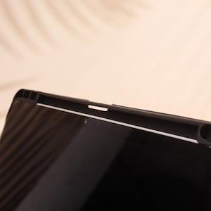 May include: Close-up of a black tablet with a protective case. The case has a textured design along the top edge and a white accent line. The screen is black and reflective, with a small camera lens visible.