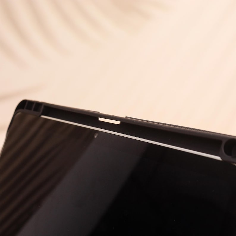 May include: Close-up of a black tablet with a protective case. The case has a textured design along the top edge and a white accent line. The screen is black and reflective, with a small camera lens visible.