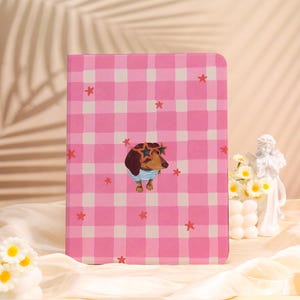 May include: A pink and white checkered notebook with a dachshund wearing star-shaped sunglasses and a striped shirt. Red star accents are scattered across the cover. The notebook is a rectangular shape.