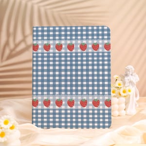 May include: A blue and white gingham pattern notebook with a border of red strawberries. The notebook is rectangular and has a soft, fabric-like texture. The background is a light beige.