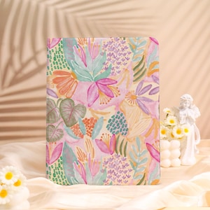 Könnte beinhalten: Ein Hardcover-Notizbuch mit einem lebendigen Aquarell-Blumenmuster. Das Design zeigt rosa, orange und grüne Blumen und Blätter auf einem hellrosa Hintergrund. Das Notizbuch ist rechteckig und scheint geschlossen zu sein.