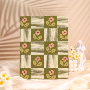 May include: A floral patterned notebook with a green and white checkered design. The notebook features pink flowers and green leaves. The background is a soft, neutral color.