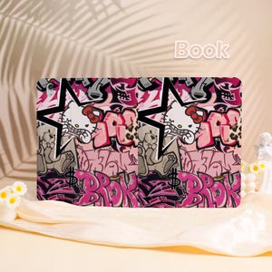 May include: A tablet case with a pink and black graffiti design. The case features a cartoon cat character, stars, and the word "BROK" in pink. The word "Book" is written in white at the top. The design is repeated across the case.