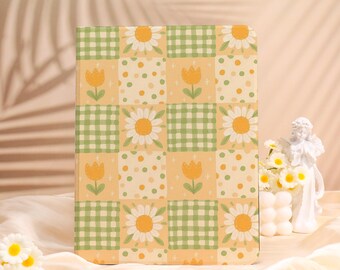 May include: A rectangular notebook with a patchwork design featuring daisies, tulips, polka dots, and green gingham squares on a yellow background. The notebook is standing upright on a cream-colored surface.