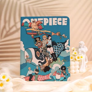Könnte beinhalten: Ein blaues Notizbuch mit einer farbenfrohen Illustration von Charakteren aus der Anime-Serie "One Piece". Das Cover zeigt eine Gruppe von Figuren, die auf einem stilisierten Fisch vor blauem Himmel reiten. Das Wort "ONE PIECE" ist oben aufgedruckt.