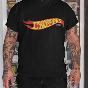 Puede incluir: Camiseta negra con un logotipo "Choppers" rojo y amarillo en forma de llama. El logotipo es un logotipo Hot Wheels estilizado.
