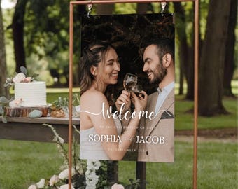 May include: A wedding welcome sign with a photograph of a smiling couple holding wine glasses. The sign reads "Welcome" and displays the names "Sophia and Jacob" with the date "25. September 20XX". The sign is in a copper frame.
