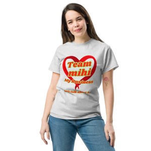 Pode incluir: Camiseta cinza claro com um design em forma de coração vermelho com as palavras douradas "Team mihi". Abaixo do coração, o texto "My Business" e "Let's talk about it" são visíveis. A camiseta é usada com jeans azul.