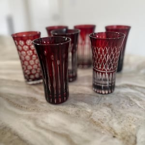 Puede incluir: Seis vasos de chupito de vidrio rojo con intrincados diseños de vidrio cortado. Los vasos están dispuestos en semicírculo sobre una superficie de mármol blanco.