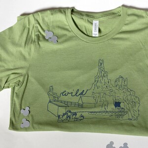 May include: A light green t-shirt with a blue graphic of a mountain range and the word "wild". The shirt is made of a soft material and is folded. Gray Mickey Mouse-shaped confetti is scattered around the shirt.