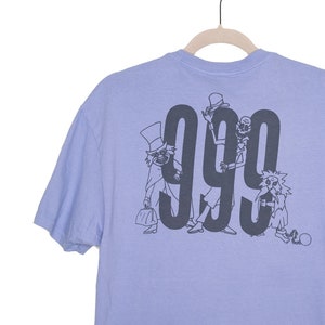 Può includere: T-shirt lilla chiaro con una grafica grigio scuro del numero "999" e personaggi di fantasmi dei cartoni animati. La t-shirt è appesa a un appendiabiti di legno.