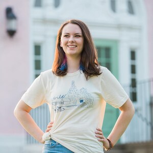 May include: A woman wearing a cream-colored t-shirt with a blue graphic and the word "wild". The shirt is paired with blue jeans. The background features a pink building with a white arched doorway and a green door.