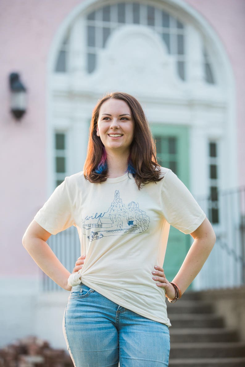 May include: A woman wearing a cream-colored t-shirt with a blue graphic and the word "wild". The shirt is paired with blue jeans. The background features a pink building with a white arched doorway and a green door.
