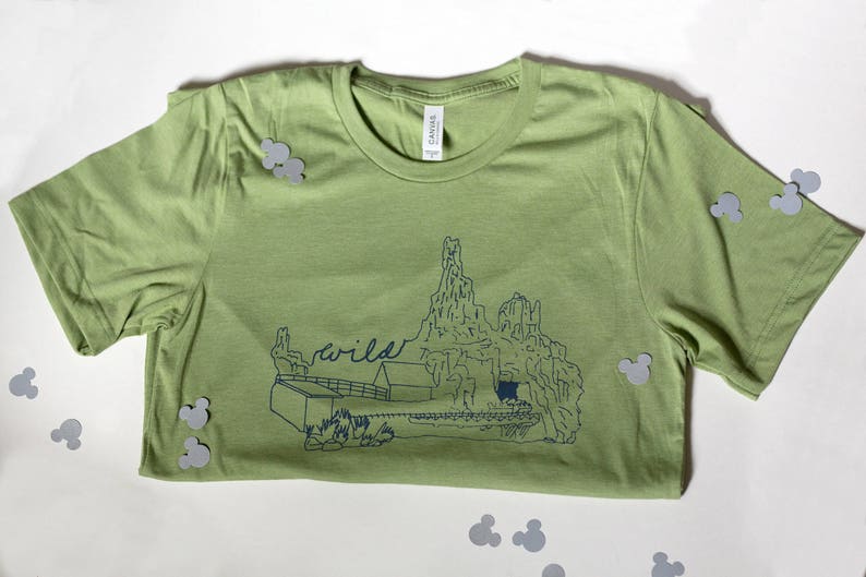 May include: A light green t-shirt with a blue graphic of a mountain range and the word "wild". The shirt is made of a soft material and is folded. Gray Mickey Mouse-shaped confetti is scattered around the shirt.