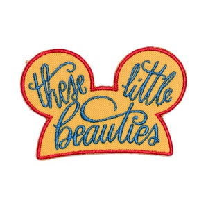 May include: Embroidered patch in the shape of Mickey Mouse ears, with the words "these little beauties" in blue script on a yellow background, outlined in red.