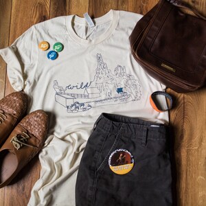 May include: An assortment of clothing and accessories, including a cream-colored t-shirt with a mountain graphic and the word "wild", dark brown leather shoes, a brown leather bag, and dark gray shorts. There are also three small round pins and an orange and black bracelet.