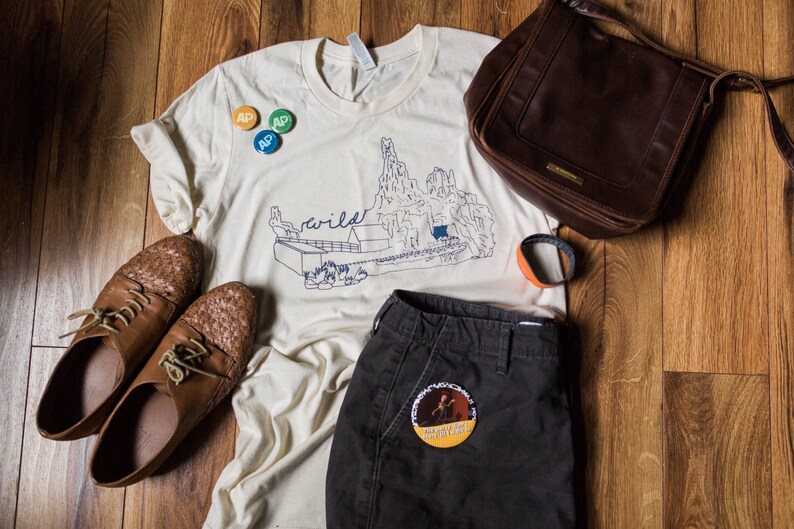 May include: An assortment of clothing and accessories, including a cream-colored t-shirt with a mountain graphic and the word "wild", dark brown leather shoes, a brown leather bag, and dark gray shorts. There are also three small round pins and an orange and black bracelet.