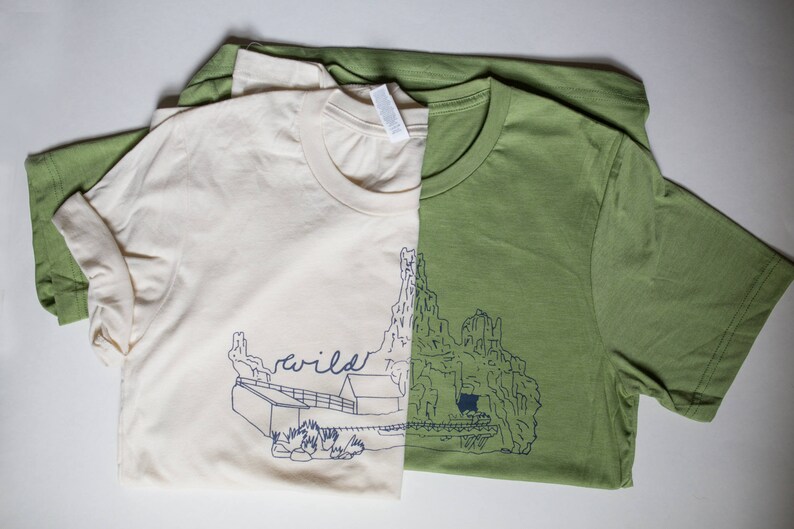 May include: Two t-shirts, one cream and one olive green, are folded and layered. The cream shirt has a blue illustration of a cabin and the word "wild". The green shirt has a blue mountain illustration.