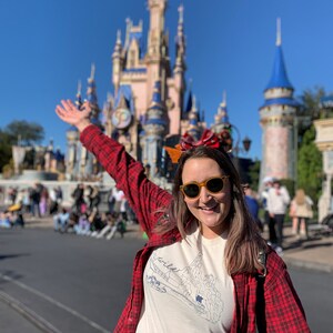 May include: A person wearing a white t-shirt with a mountain graphic and the word "wild" in blue, paired with a red and black plaid shirt and yellow sunglasses. The background features a large castle under a clear blue sky.